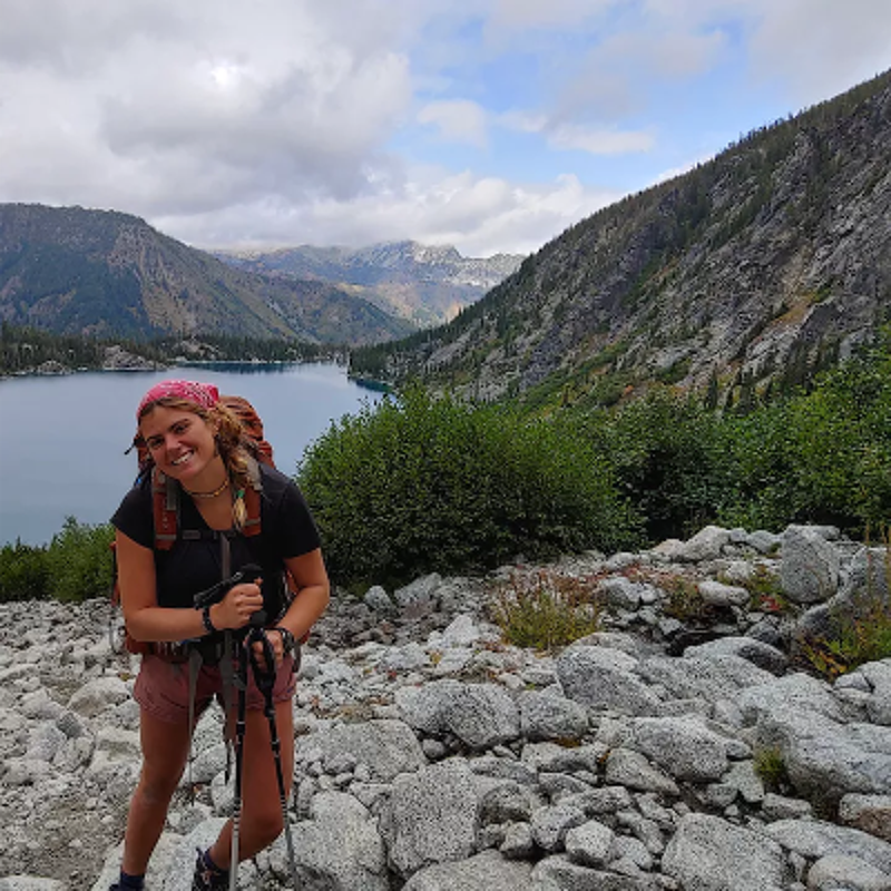 The image shows a young woman hiking in a scenic mountain landscape. She is smiling and wearing a backpack, hiking poles, and a bandana. In the background, there is a lake surrounded by mountains and a cloudy sky. The overall impression is one of adventure and natural beauty.